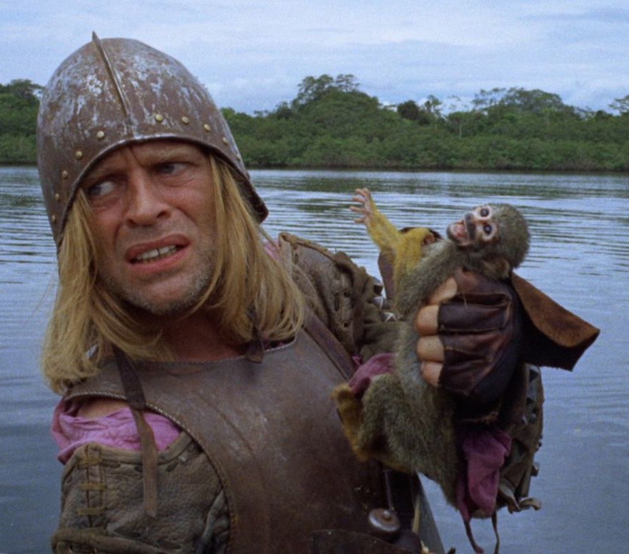 Movie still. A man in conquistador armor holds a small monkey and looks out of frame with a pained and confused expression