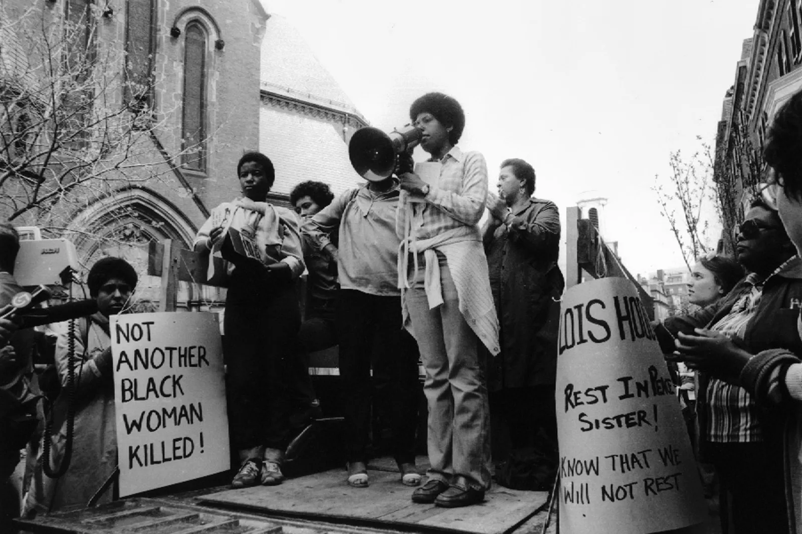Black and white photo. Small group of Black women stand on a small platform. One holds a megaphone to her mouth. Another holds a sign by her feet that reads 'NOT ANOTHER BLACK WOMAN KILLED!