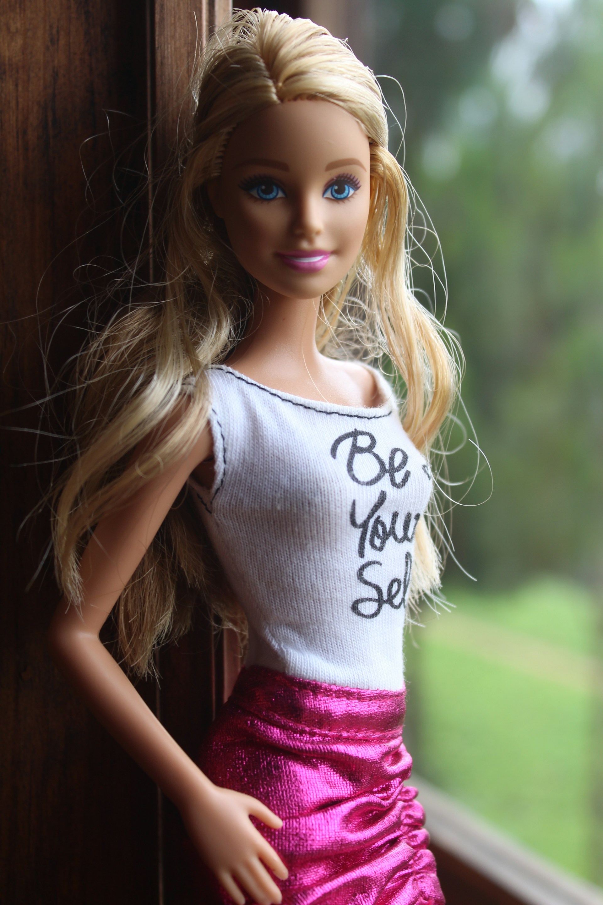 Close-up hoto of a Barbie doll posed in front of a window wearing a tanktop with the words 'Be Your Self'