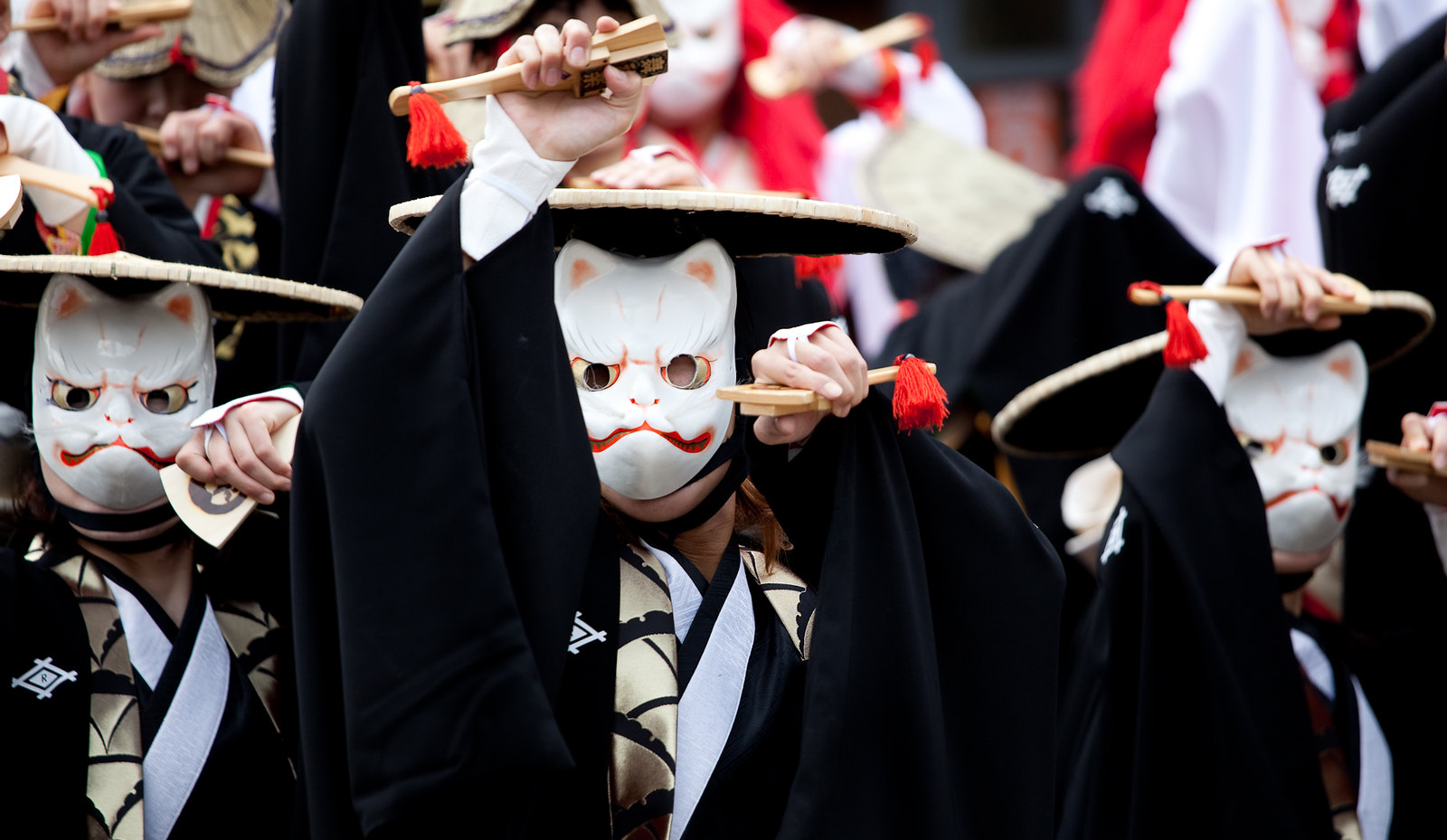 Several people in a parade, wearing Japanese style costumes and white cat masks. They appear to doing a coreographed dance.