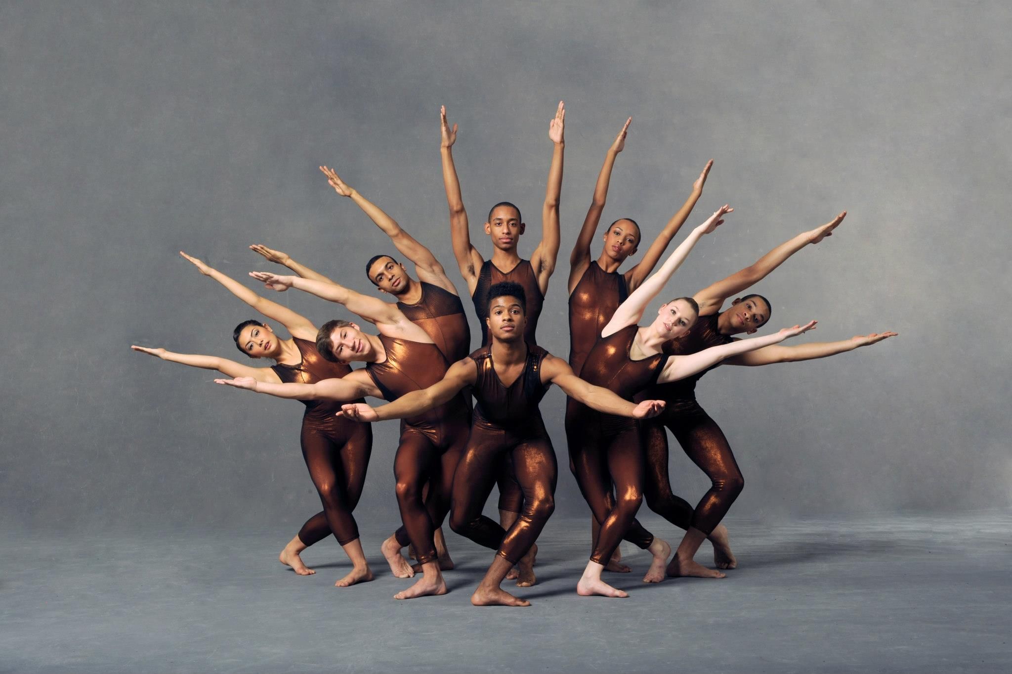 A troupe of dancers in leotards with their arms and bodies positioned to make a radial pattern with their outstretched hands