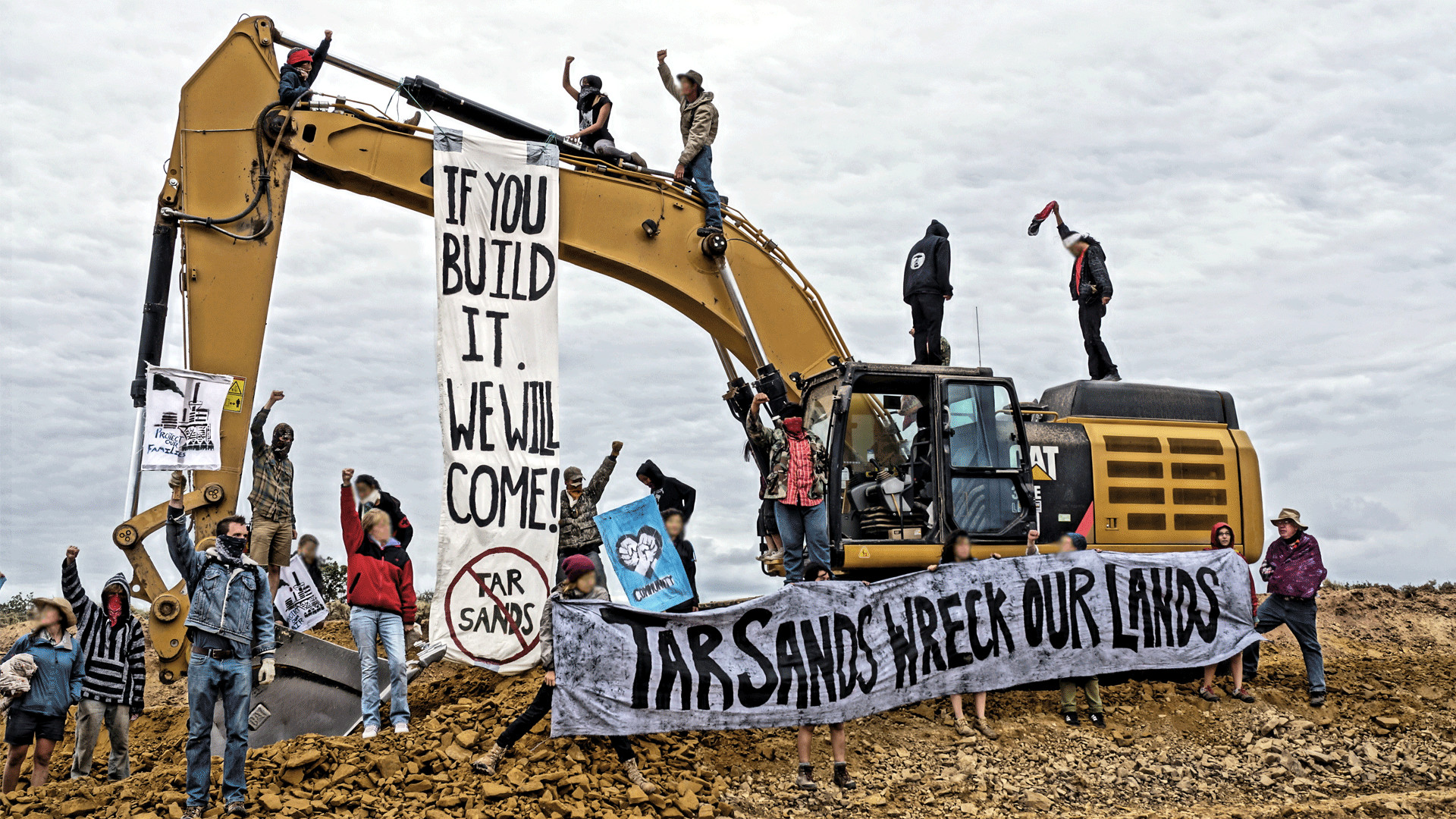 Photo of protesters surrounding and on top of a large backhoe, most of their fists raised above their heads. A large banner hangs from the arm of the backhoe reading 'If you build it we will come' and 'Tar sands' with a red cross through it. A second banner is held by protesters on the ground, reading 'tar sands wreck our lands'.