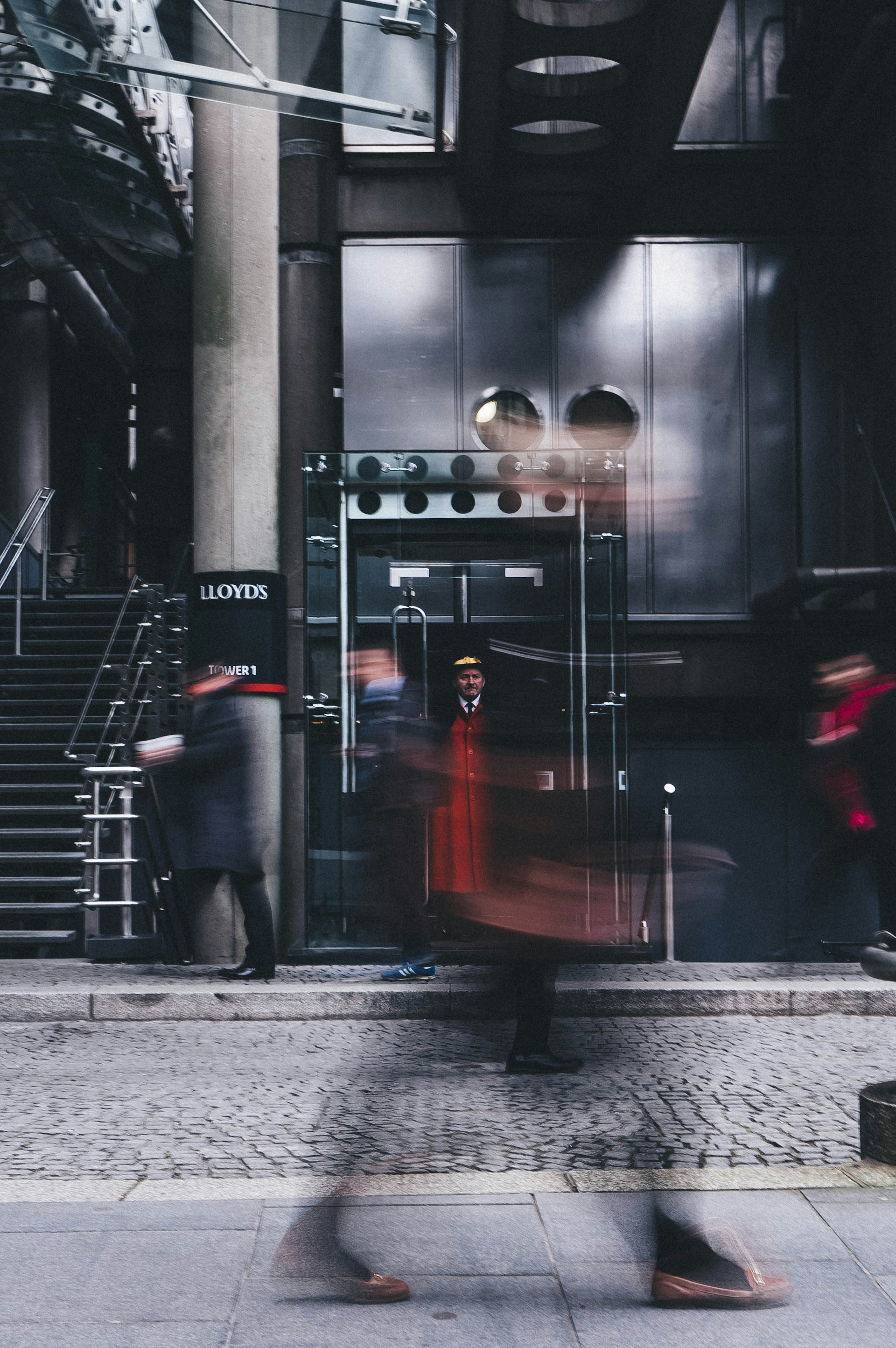 Photo of a doorman standing still in front of Lloyds of London with blurred images of other people walking quickly by.