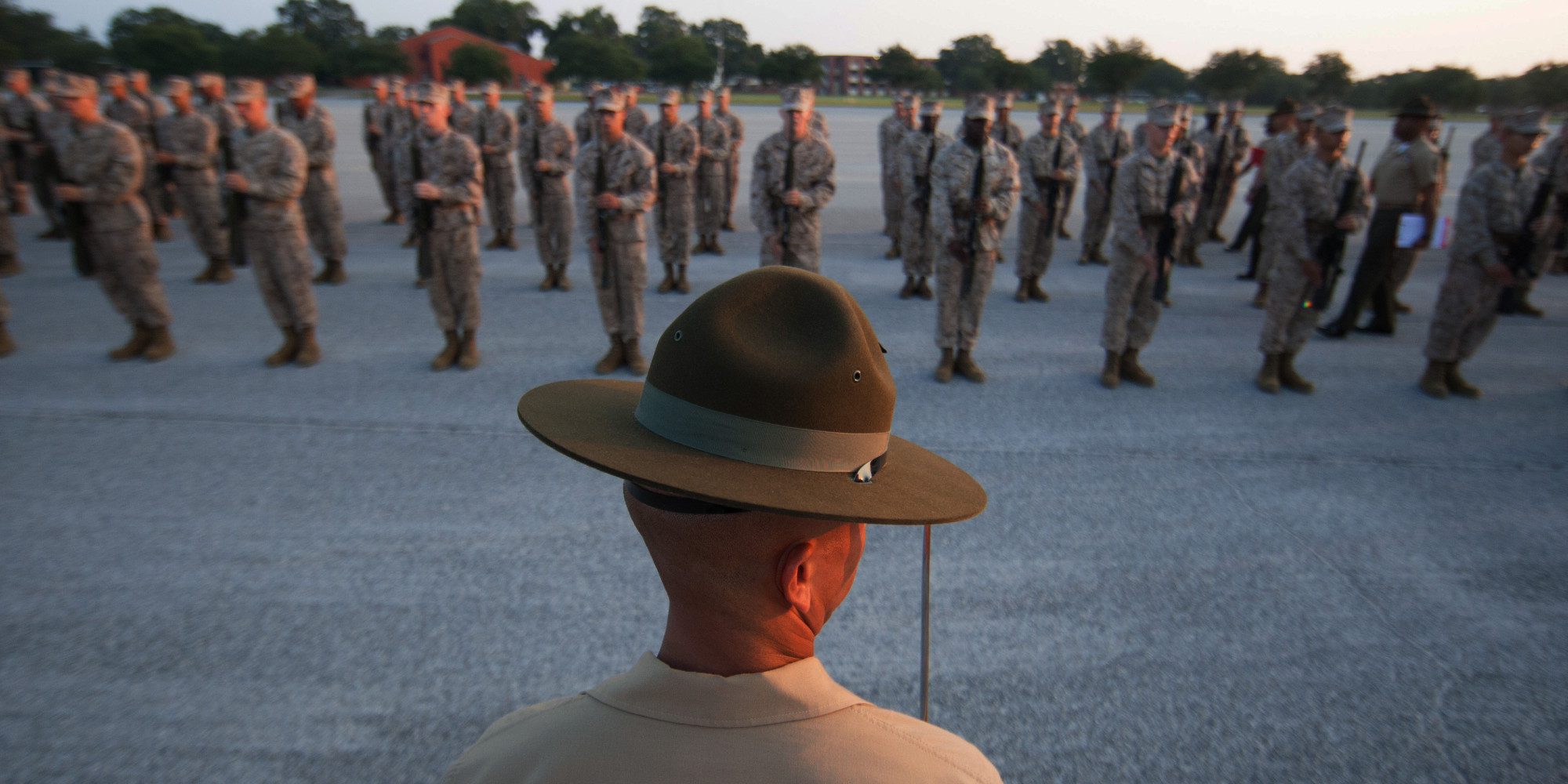 Foreground: a military drill sergean from behind, looking at the background. Background: an orderly group of soldiers in fatigues holding guns