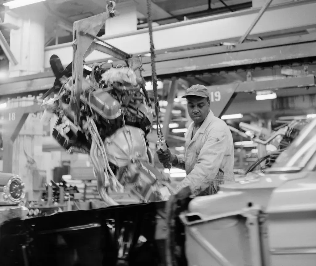 Black and white photo. A Black man in an auto factory lowers an engine into a car.
