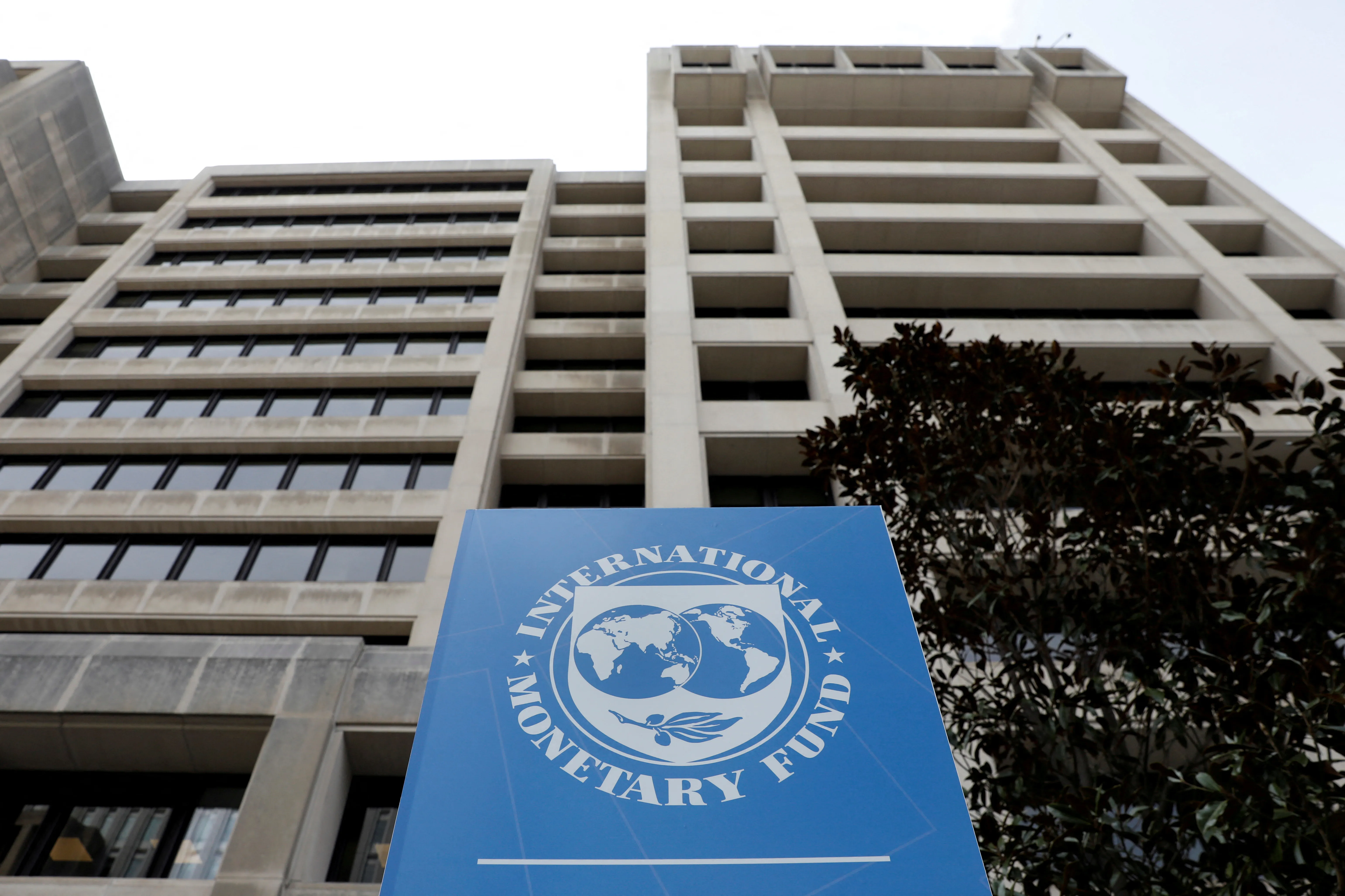 Photograph looking up at a tall generic office building. A blue sign is in front of the building with the logo fo the International Monetary Fund: a Shield with a map of the Earth and an olive branch, with the words 'International Monetary Fund' in a circle around it.