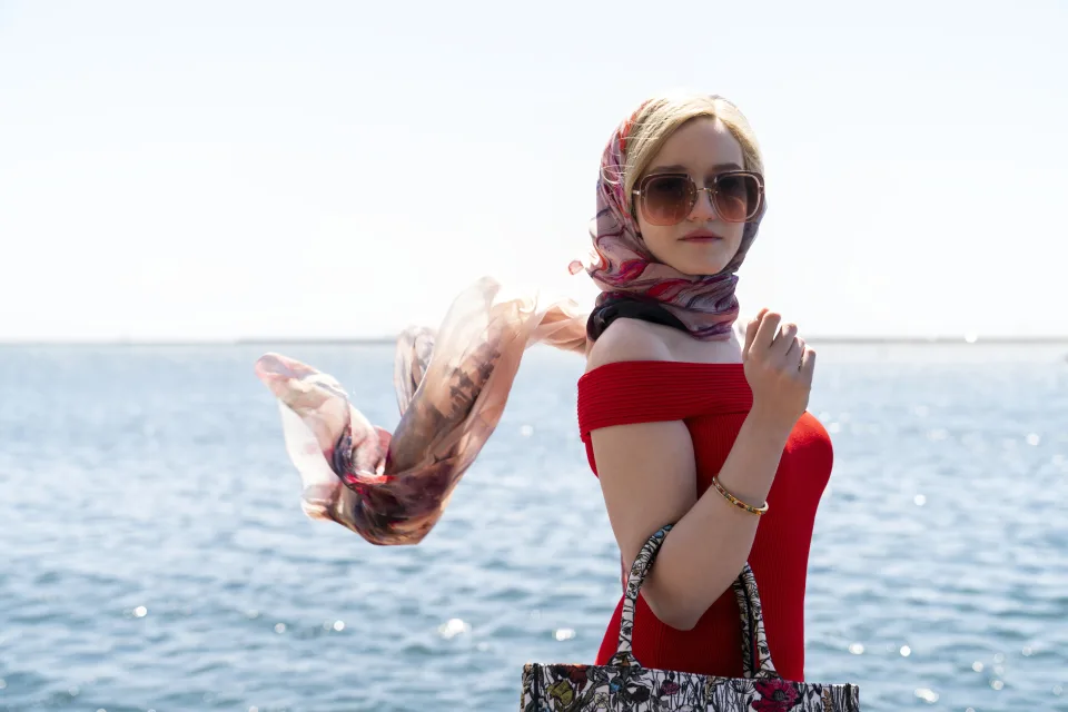 Photo of a woman standing by the seaside wearing expensive clothing and large sunglasses. Her scarf blows behind her in the wind