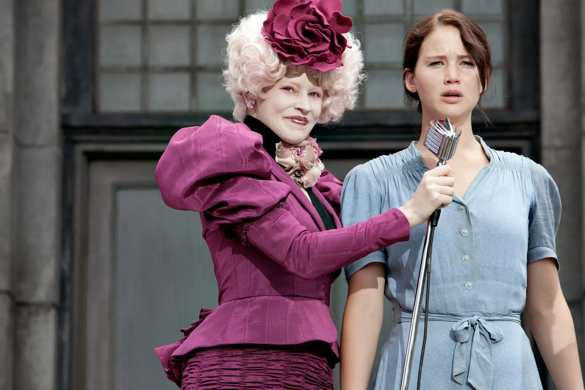 Screenshot from The Hunger Games. A plainly dressed woman (Katniss) stands next to an extravagantly dressed woman (Effie Trinket) in front of a microphone