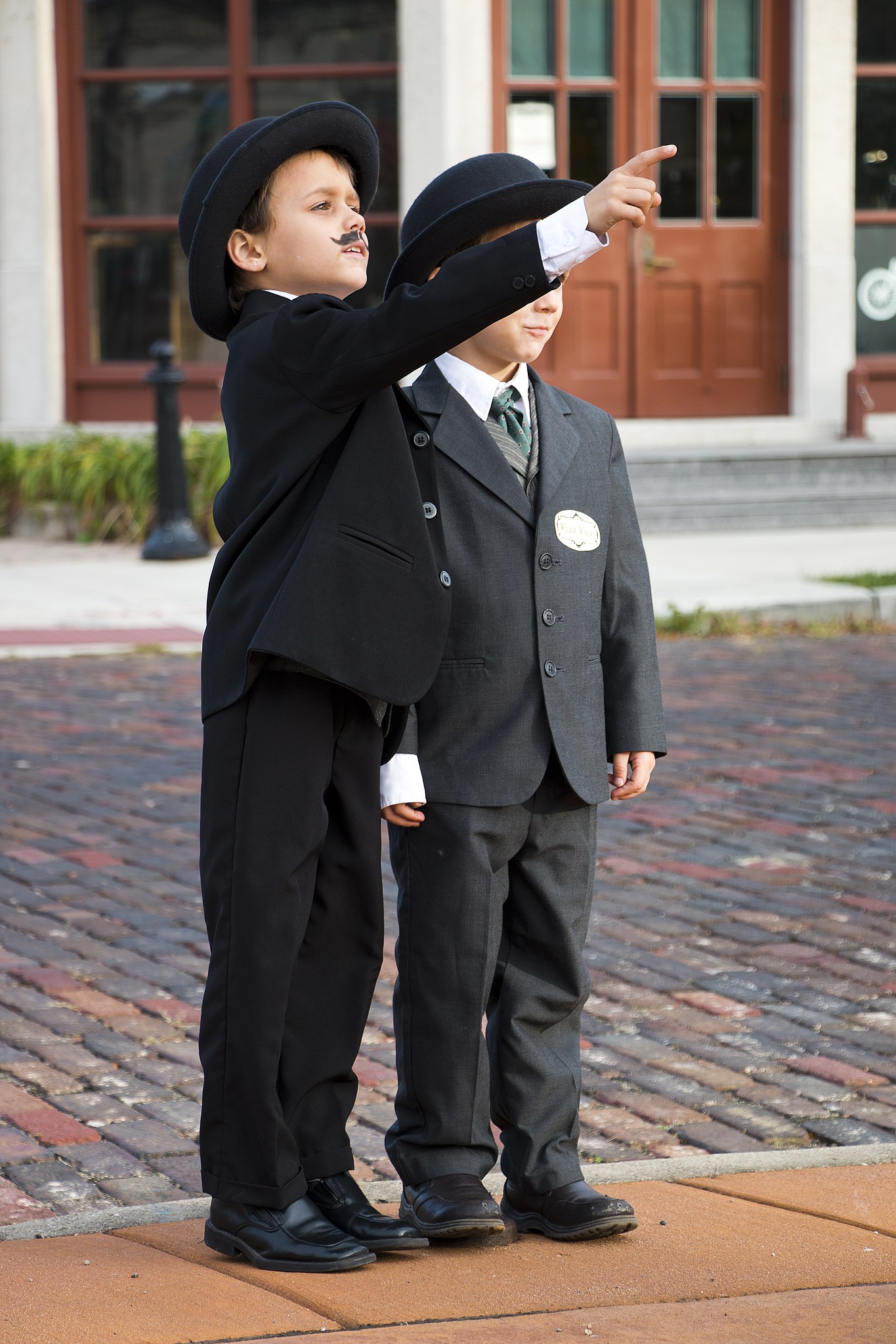 Two kids wearing 19th-century style suits. One of them is pointing toward the horizon.