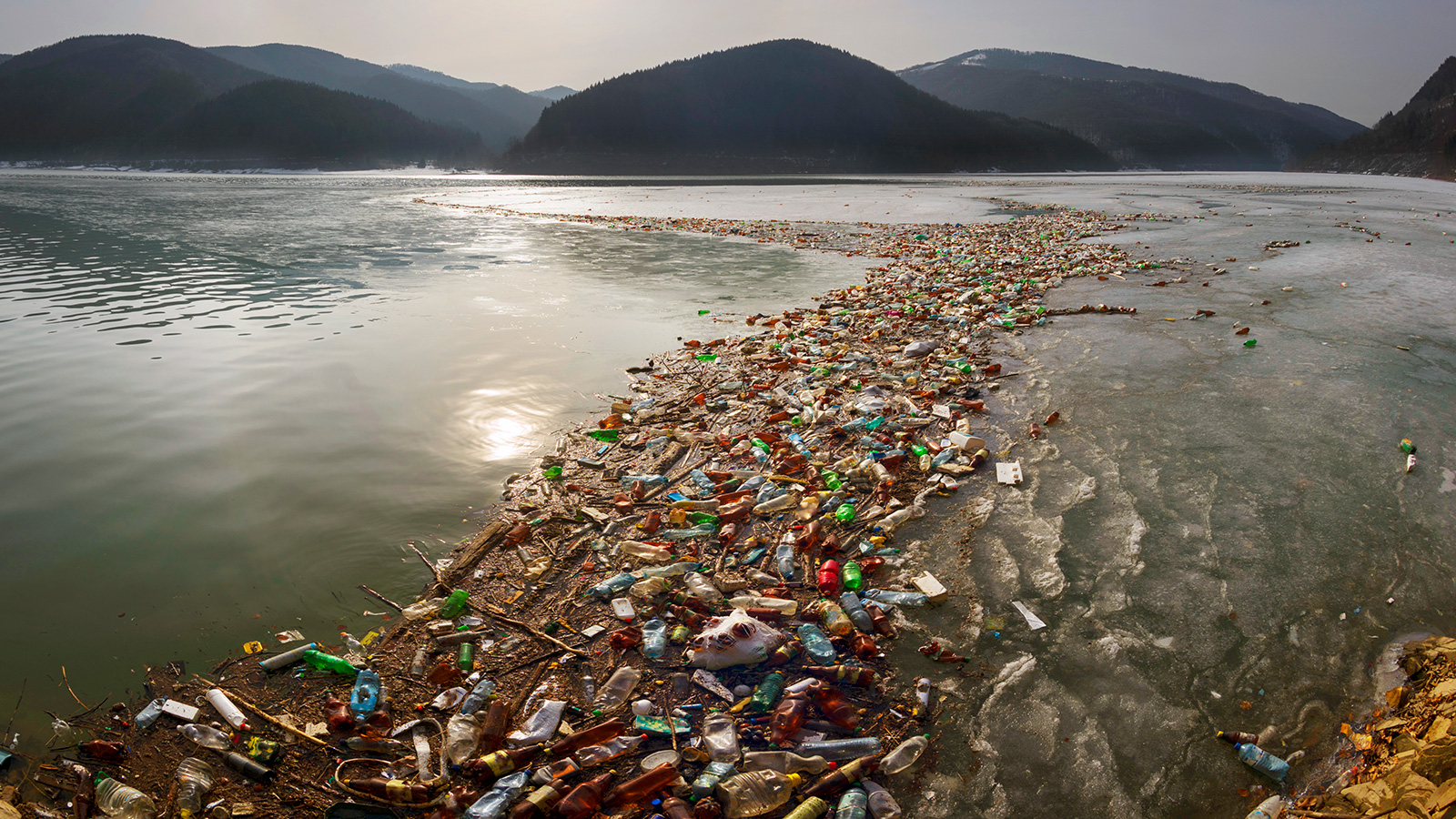 Photo of an ocean bay with mountains in the background. A floating river of colorful plastic garbage flows into the bay.