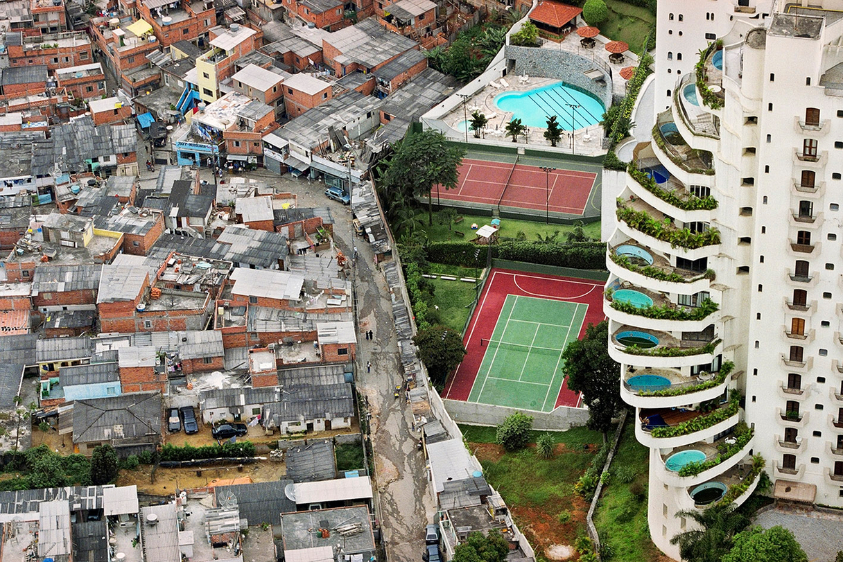Aerial photo of an urban area with a clear division down the middle. On the left there are many small buildings and houses, many in run-down condition. On the right there is a large, luxurious condo complex or hotel, including very big tennis courts ands a swimming pool on each individual balcony.