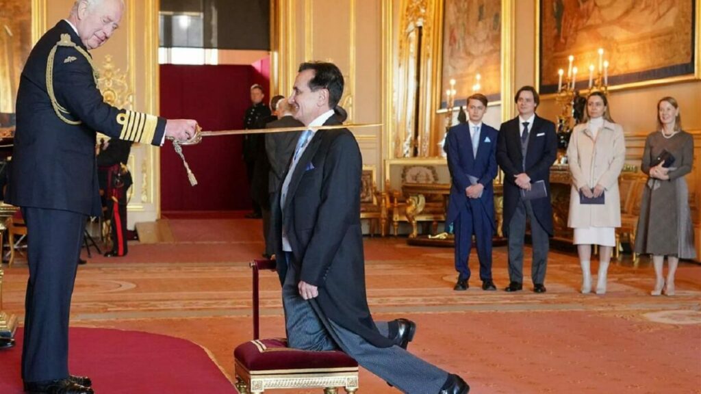 Photograph of King Charles of England tapping a kneeling man in a suit on the shoulder with a ceremonial sword. Four people stand in the background watching. (knighting ceremony)