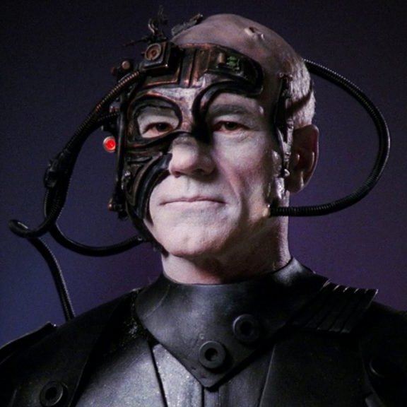 Photo of the head and shoulders human man with completely white skin covered in electronics, including an eyepiece over his right eye that glows red. (Captain picard from Star Trek as a member of the Borg)