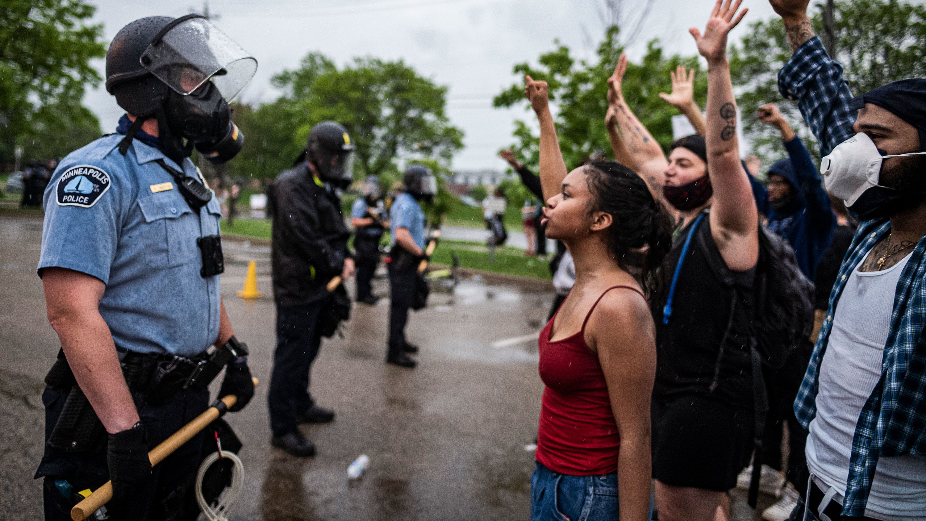 Photograph of several protesters with arms raised confonting a line of police wearing riot gear