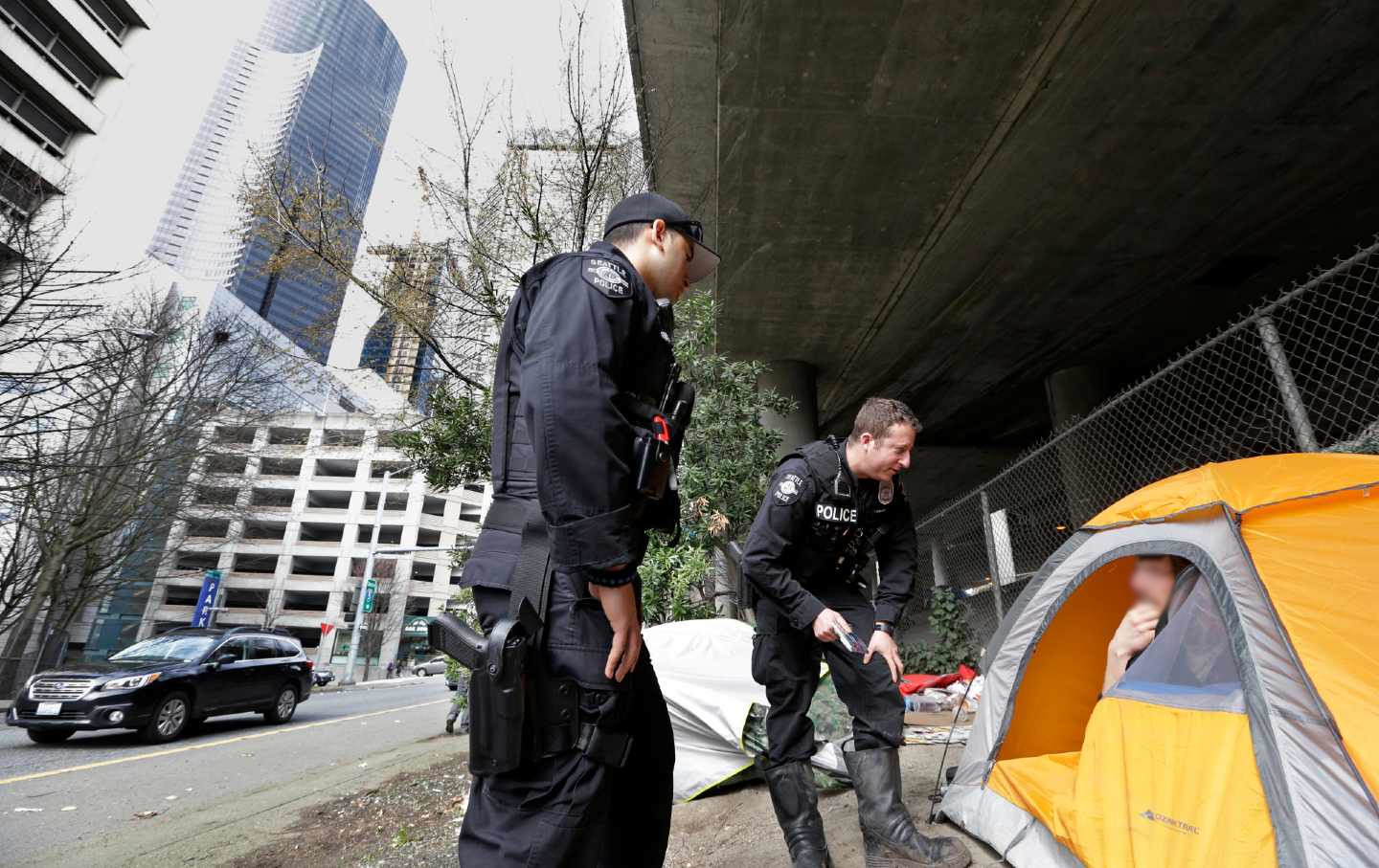 A cop leans over to talk to a person inside a yellow tent under a highway overpass.