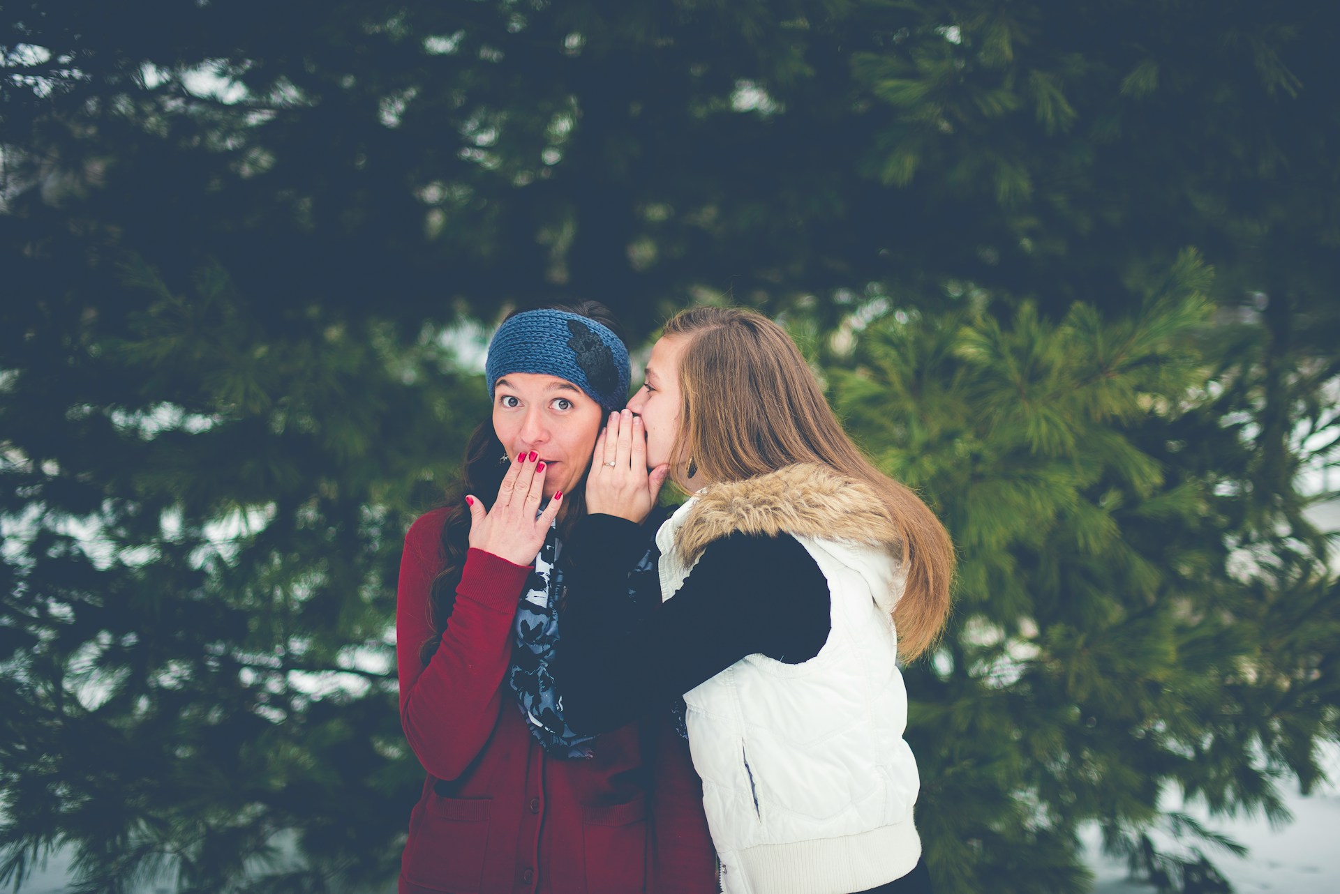 Photo of two people in  the woods. The person on the right is whispering into the person on the left's ear. The person on the left is looking at the camera and covering their mouth in a feigned expression of scandalous surprise.
