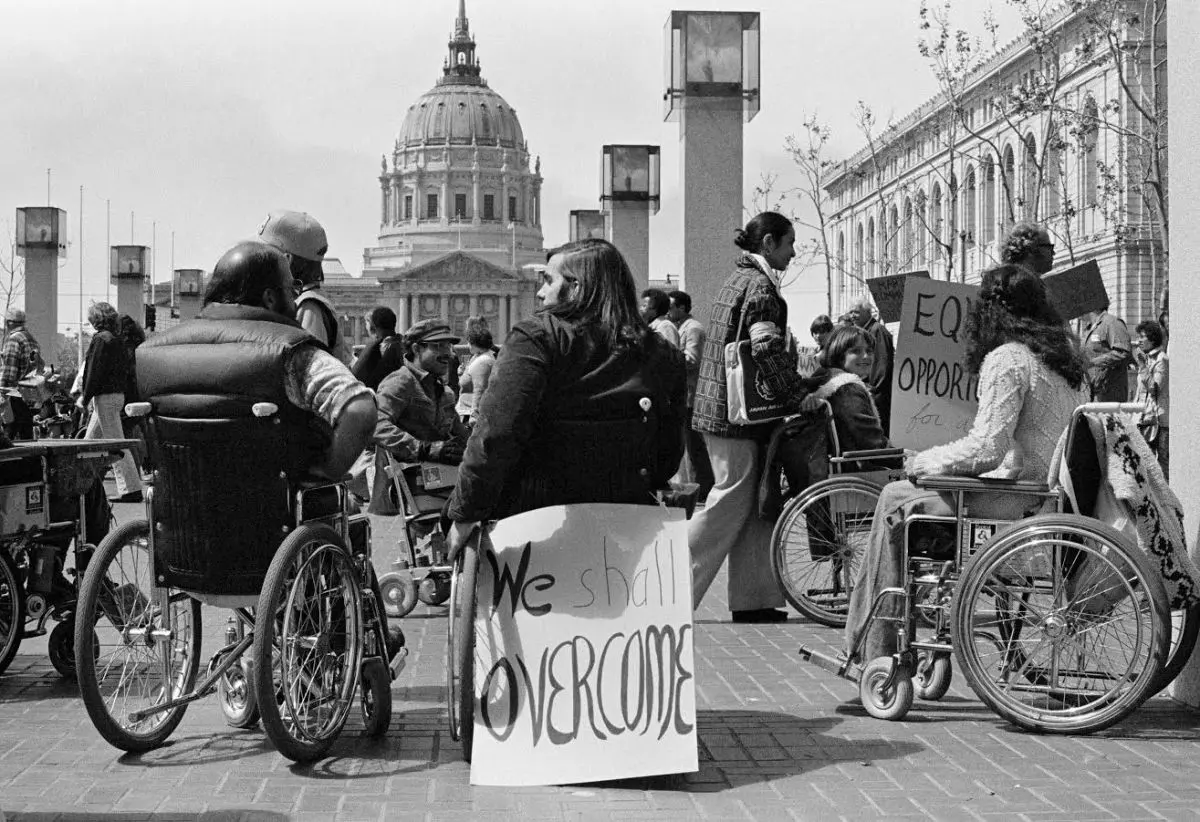 Black and White photo of a crowd of people, mostly in wheelchairs, in front of a capital building. The closest wheelchair to the camera has a sign attached reading 'We shall overcome'