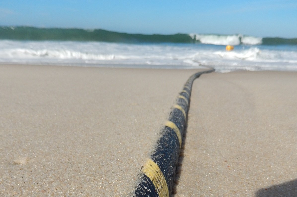 Photograph of a black cable with yellow spiral stripe on a sandy beach. In the background, the cable descends into the ocean.