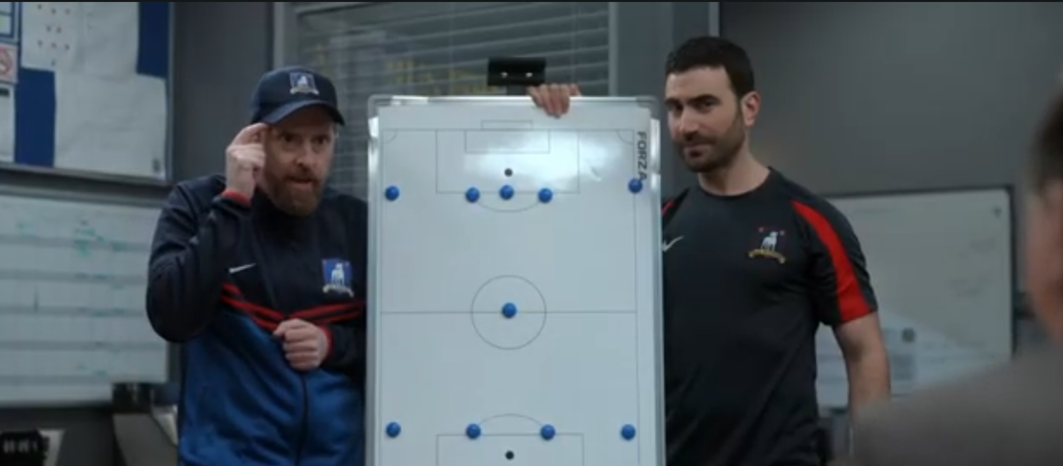 Two men standing on either side of a whiteboard with a soccer field. Blue magnets show player positions on the board. They are explaining something to a third person, barely visible in the foreground. One of the men is tapping his index finger against his head.