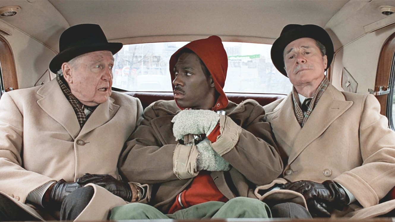Still from Trading Places. A shabbily dressed Black man (Eddie Murphy) sits between two older white men in matching expensive coats, hats, and gloves in the back of a limosine