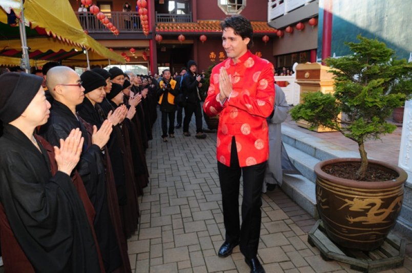 Justin Trudeau wearing a Chinese style shirt, with hands pressed palms together in a Buddhist temple. A line of people are making the same hand gesture to him.