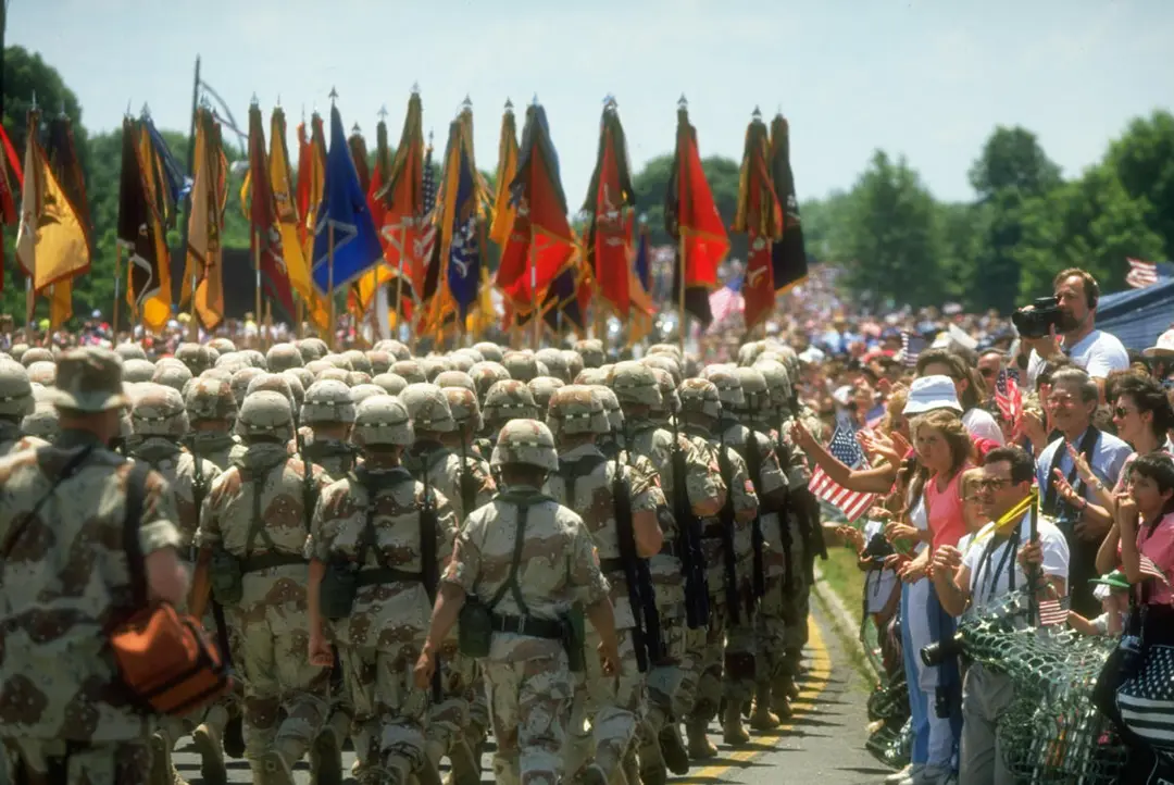 A military parade of camo-clad soldiers carries an array of inscrutible flages as it moves away from the camera. A large crowd of spectators look on, some holding United States flags.