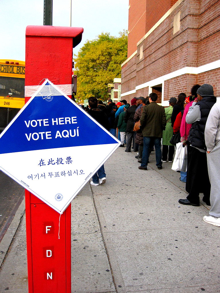 A line of people with their backs to the camera on a sidewalk. In the foreground, a diamond-shaped sign reads 'Vote Here' in four languages.