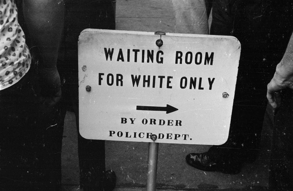 Black and white photo of a sign reading 'WAITING ROOM FOR WHITE ONLY; BY ORDER POLICE DEPT' with an arrow pointing to the right.